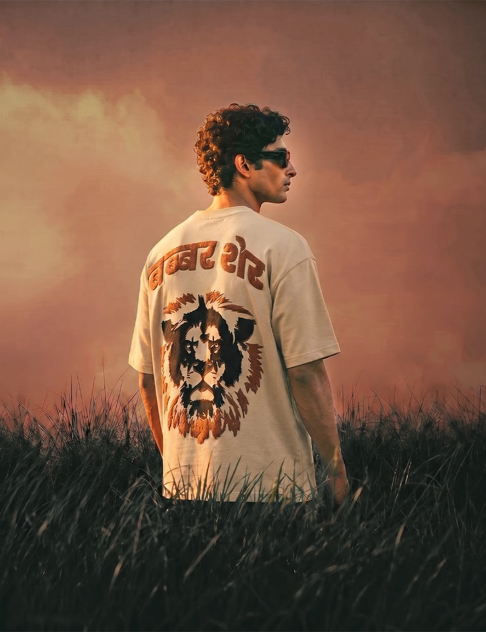 Model in Naram Babbar Sher beige oversized graphic tee standing in grass field at golden hour, Hindi lion back print visible, streetwear lifestyle