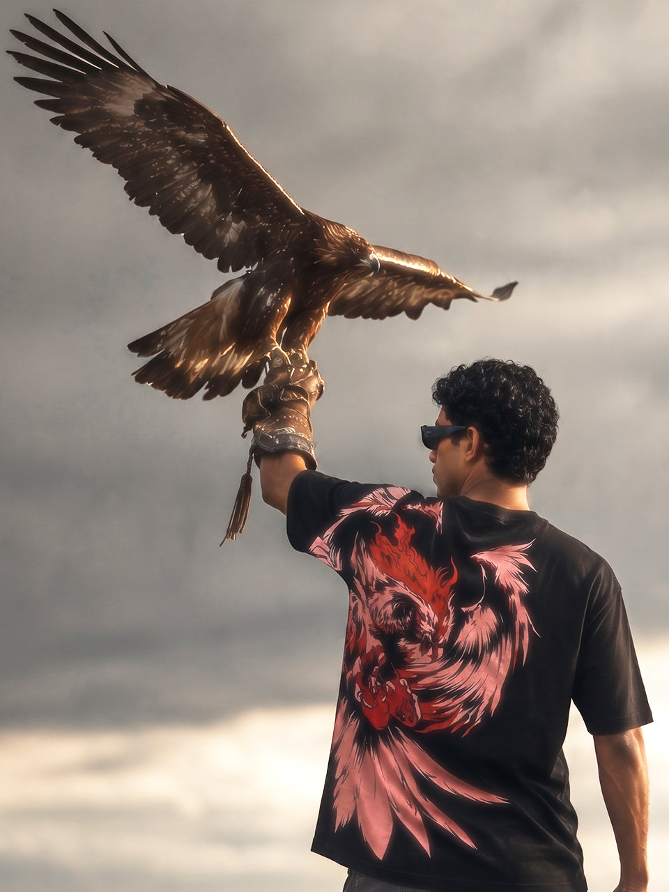 
Model wearing Naram Eagle black oversized graphic tee holding real eagle on arm against stormy sky, Indian streetwear