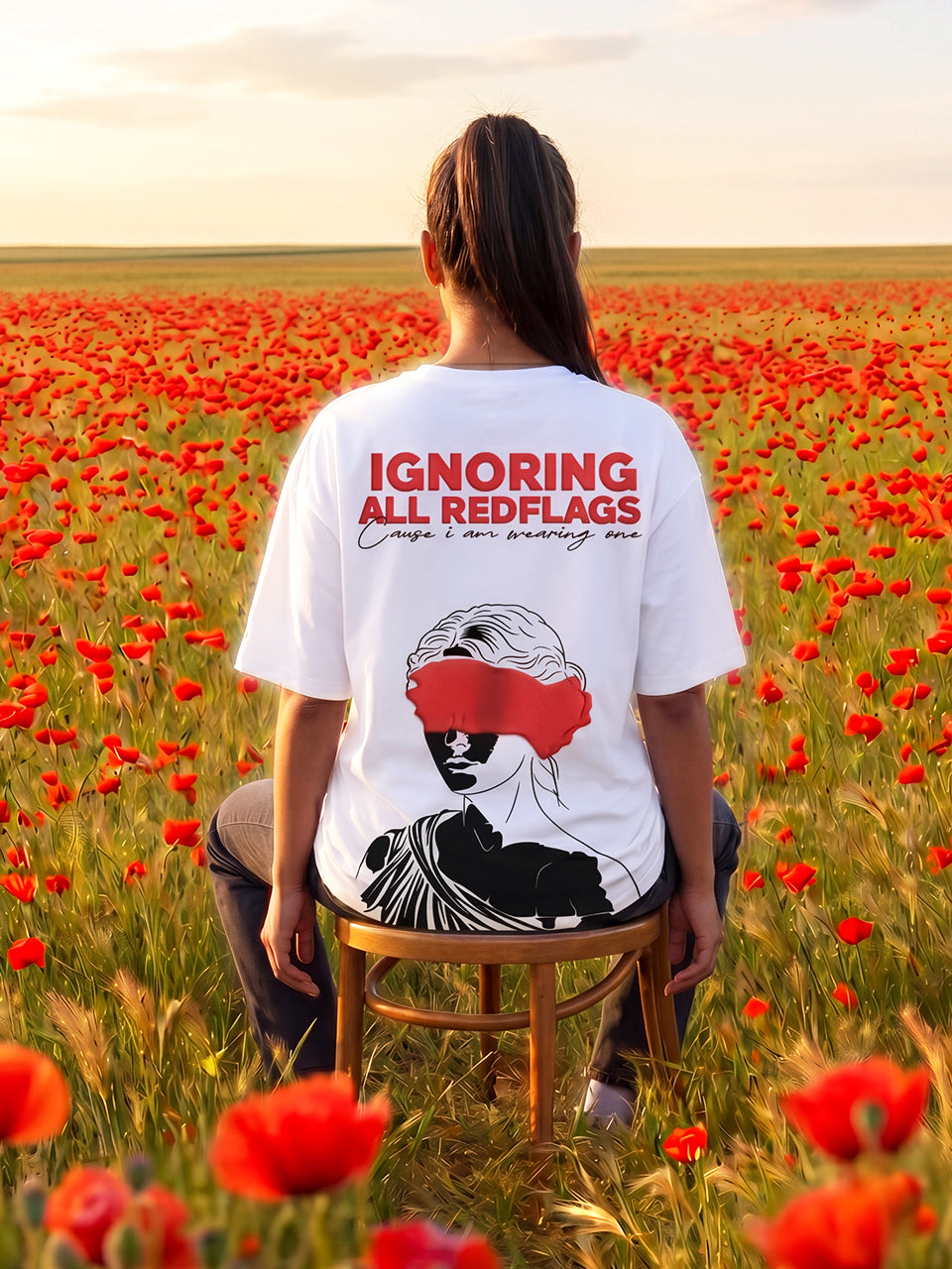 Female model in Naram white oversized graphic tee seated on wooden chair in red poppy field showing Red Flag back print, women streetwear lifestyle India