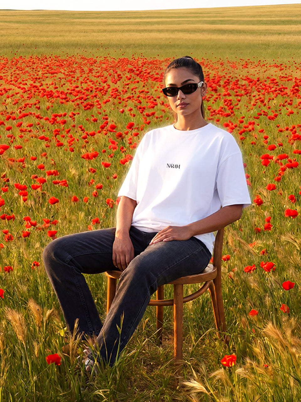 Female model wearing Naram white oversized tshirt seated in poppy flower field, Naram chest logo visible, Indian women streetwear lifestyle shot